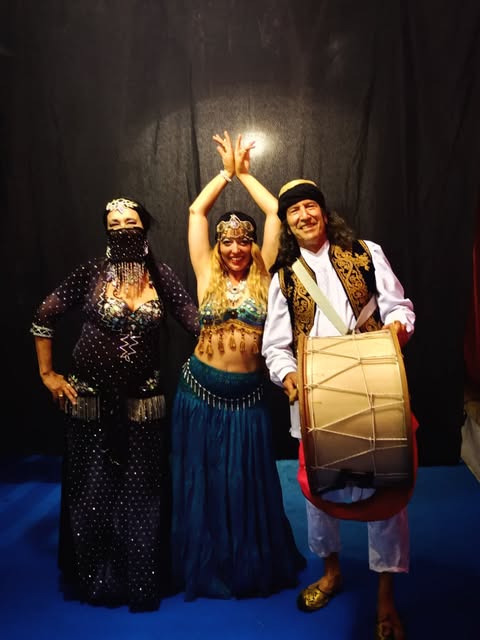 Grupo de artistas con trajes de danza ornamentados ante un fondo oscuro.