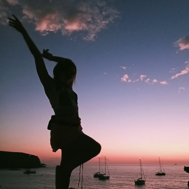 Silueta de una bailarina ante un atardecer rosa sobre una bahía, con una rodilla flexionada y los brazos elevados.